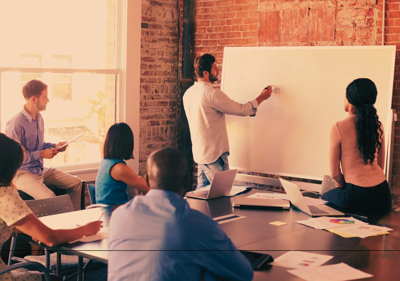 “A diverse group of professionals engaged in a brainstorming session, with a man writing on a large whiteboard in a modern brick-walled office space.”